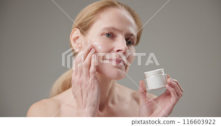 Real people - age, beauty, health and dry skin care concept - beautiful mature Caucasian middle aged woman in her 50s applying moisturizer to her face 116603922