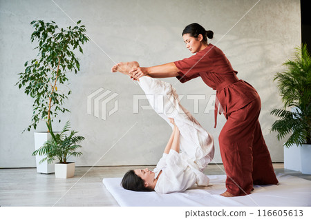 Masseuse making thai yoga massage. Woman therapist doing traditional massage treatment, stretching female patient's body. Client lying on mat, enjoying hand massage in calm atmosphere. 116605613