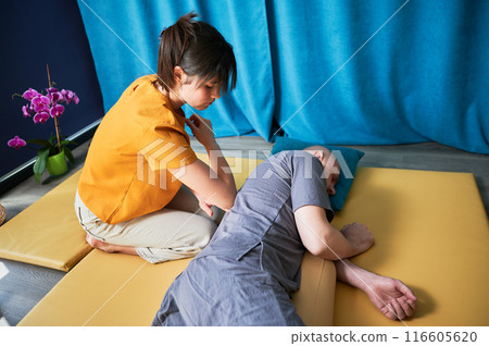 Masseuse making thai yoga massage. Woman therapist doing traditional massage treatment using elbow for male patient indoors. Client lying on mat, meditating, enjoying hand massage in calm atmosphere. Masseuse making thai yoga massage. Woman therapist doing traditional massage treatment using elbow for male patient indoors. Client lying on mat, meditating, enjoying hand massage in calm atmosphere. 116605620