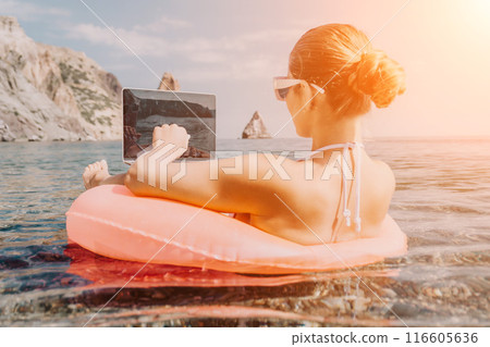 Woman freelancer works on laptop swimming in sea on pink inflatable ring. Happy tourist in sunglasses floating on inflatable donut and working on laptop computer in calm ocean. Remote working anywhere Woman freelancer works on laptop swimming in sea on pink inflatable ring. Happy tourist in sunglasses floating on inflatable donut and working on laptop computer in calm ocean. Remote working anywhere 116605636