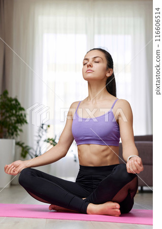 Caucasian woman with closed eyes practicing yoga and meditation sitting in lotus pose on pink yoga mat at home Caucasian woman with closed eyes practicing yoga and meditation sitting in lotus pose on pink yoga mat at home 116606514