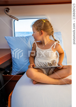 Little girl sits with tablet in the cabin of a yacht and looks out the window during a sea crossing. Yacht life concept 116606523