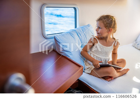 Little girl sits with tablet in the cabin of a yacht and looks out the window during a sea crossing. Yacht life concept Little girl sits with tablet in the cabin of a yacht and looks out the window during a sea crossing. Yacht life concept 116606526