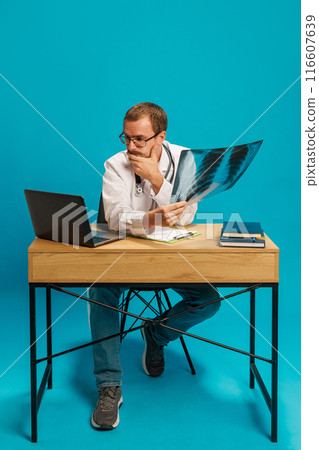 Man, doctor sitting at table, holding x ray and looking on laptop with thoughtful face against cyan background Man, doctor sitting at table, holding x ray and looking on laptop with thoughtful face against cyan background 116607639