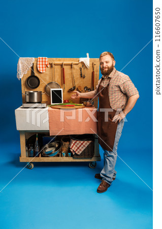 Chef, bearded man in casual attire, wearing an apron, pointing at mobile kitchen setup, with pot, vegetables and tablet on table against blue background Chef, bearded man in casual attire, wearing an apron, pointing at mobile kitchen setup, with pot, vegetables and tablet on table against blue background 116607650