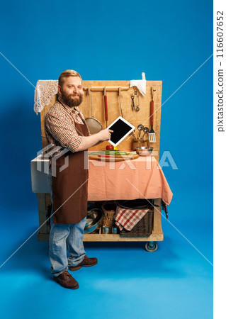 Smiling bearded man in plaid shirt and apron, standing next to small kitchen, pointing on tablet screen against blue background Smiling bearded man in plaid shirt and apron, standing next to small kitchen, pointing on tablet screen against blue background 116607652
