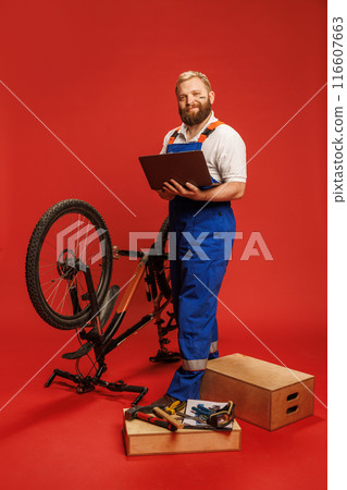 Bearded smiling young man in blue overalls, mechanic standing with laptop next to bicycle against red background. Online services. Bearded smiling young man in blue overalls, mechanic standing with laptop next to bicycle against red background. Online services. 116607663