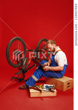 Bearded young man in blue overalls, mechanic sitting next to bicycle and looking on laptop against red background 116607664