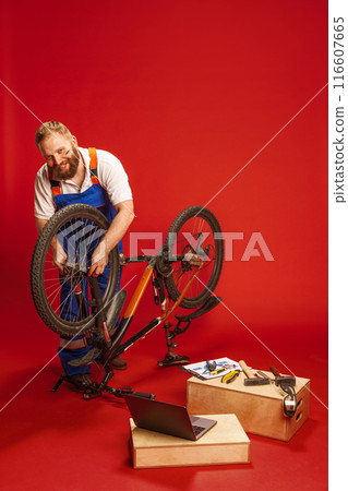 Mechanic in blue overalls, bearded man working on bicycle, surrounded by various tools and laptop against red background Mechanic in blue overalls, bearded man working on bicycle, surrounded by various tools and laptop against red background 116607665