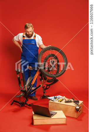 Serious bearded young man in blue overalls repairing bicycle in workshop setting, with tools and laptop on wooden boxes against red background 116607666