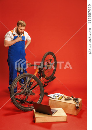 Bearded man, mechanic in blue overalls, with dirty face standing and looking on bike with thoughtful face, with laptop and tools around, against red background Bearded man, mechanic in blue overalls, with dirty face standing and looking on bike with thoughtful face, with laptop and tools around, against red background 116607669