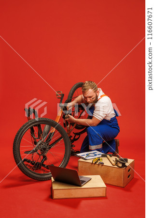 Bicycle repairman, bearded man in blue overalls working on bike, with tools and laptop on wooden boxes, against bright red background 116607671