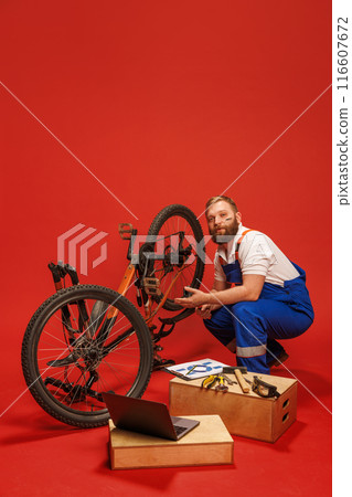 Mechanic, bearded man in blue overalls fixing bicycle, surrounded by tools and laptop, against red background Mechanic, bearded man in blue overalls fixing bicycle, surrounded by tools and laptop, against red background 116607672