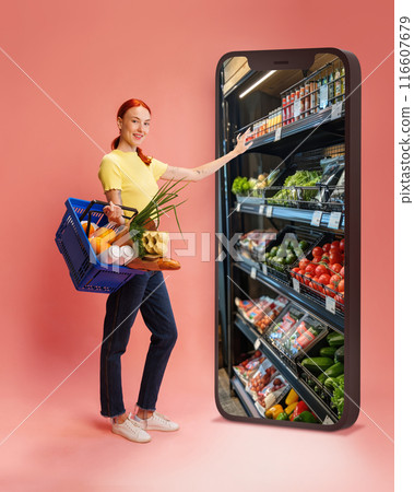 Woman in yellow shirt holding basket of groceries, reaching into giant smartphone screen displaying food shelves against pink background 116607679