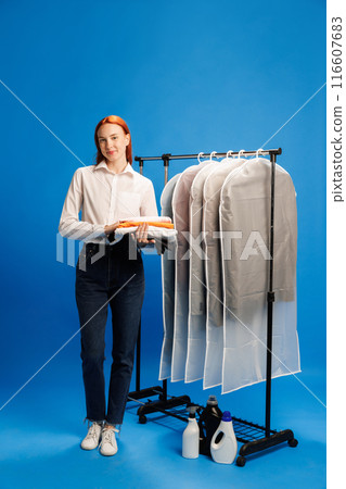 Female laundry worker in casual attire, holding stack of clothes, standing beside rack of garment bags and detergents against blue background 116607683