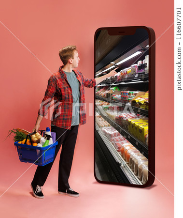 Man in red plaid shirt shopping online, holding basket of groceries, touching giant smartphone screen displaying grocery shelves against pink background 116607701