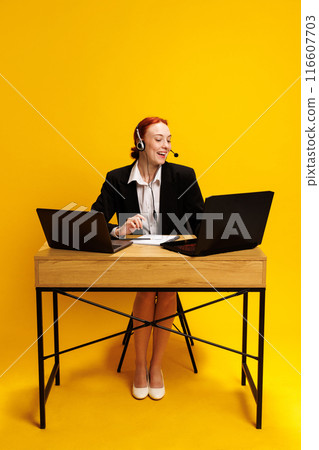 Formally dressed young woman, call center worker engaged in deep conversation, wearing headset, working at desk with two laptops against yellow background Formally dressed young woman, call center worker engaged in deep conversation, wearing headset, working at desk with two laptops against yellow background 116607703