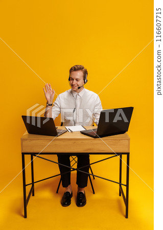 Call center agent, man in white shirt, smiling and talking on headset, seated at desk with two laptops against yellow background Call center agent, man in white shirt, smiling and talking on headset, seated at desk with two laptops against yellow background 116607715