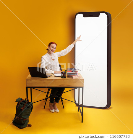 Workspace of teacher. Young man pointing at giant smartphone screen, sitting at desk with study supplies and laptop against yellow background 116607723