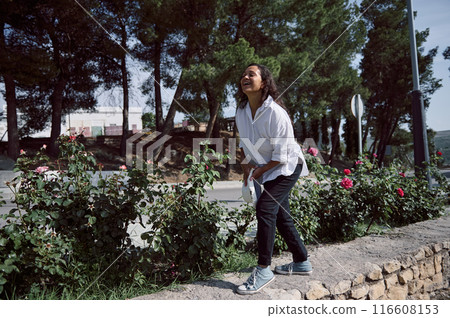 Woman enjoying nature while laughing in a sunny garden with rose bushes and tall trees Woman enjoying nature while laughing in a sunny garden with rose bushes and tall trees 116608153