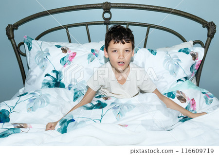 Sleepy boy with tired face is sitting in a cozy bed. 116609719