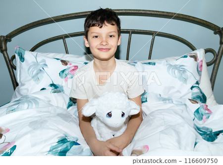 Smiling rested boy is sitting in a bed with sheep toy. 116609750