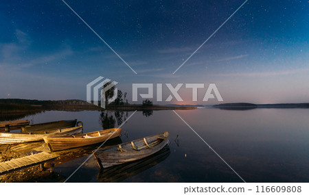 Moored Old Shabby Wooden Fishing Rowboats , Left Afloat On The Motionless Clear River Lake Water Next To Waterside. Amazing Glowing Stars Effects Above Lake. Night Starry Sky Soft Colors 116609808