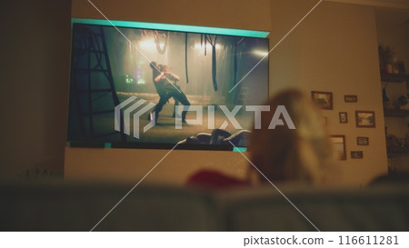 Caucasian woman sits on sofa and watches action movie on TV screen at night Caucasian woman sits on sofa and watches action movie on TV screen at night 116611281