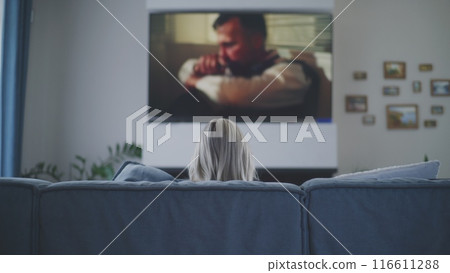Woman sits on sofa and watches action movie on TV 116611288