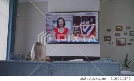 Adult woman rests on sofa in living room and watches TV news 116611545