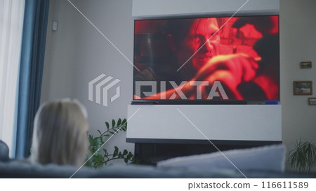 Woman sits on sofa and watches action movie on TV 116611589