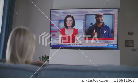 Adult woman rests on sofa in living room and watches TV news 116611608