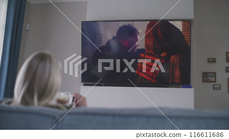 Woman sits on sofa and watches action movie on TV 116611686