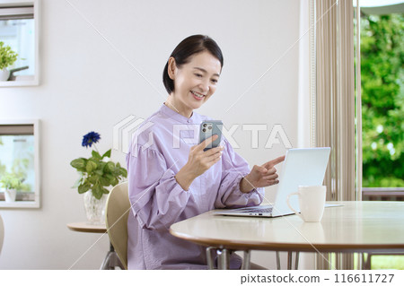 A middle-aged woman looking at a computer while using her smartphone in the living room A middle-aged woman looking at a computer while using her smartphone in the living room 116611727