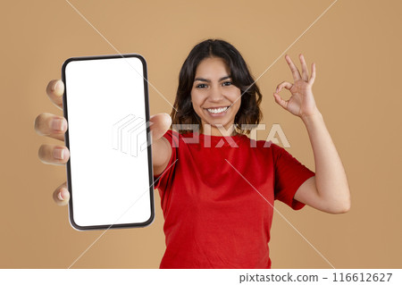 A young middle eastern woman is smiling and holding a smartphone with a blank screen in front of her. She is wearing a red shirt and gesturing the ok sign with her other hand A young middle eastern woman is smiling and holding a smartphone with a blank screen in front of her. She is wearing a red shirt and gesturing the ok sign with her other hand 116612627