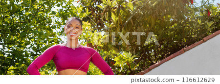 Fit young black woman exercising outdoor at garden. Portrait of confident lady training at public park, wearing pink sportswear, holding hands on waist, smiling at camera, copy space 116612629