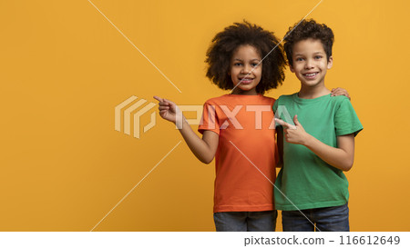 Happy African American children, a girl and a boy standing side-by-side in front of a yellow background. Both kids are smiling and pointing to the right, copy space 116612649