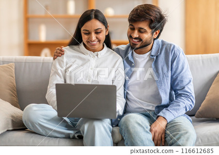 Indian man and woman are seated on a couch, both actively engaged in viewing content on a laptop screen. They appear focused and attentive, perhaps working or watching something together. 116612685