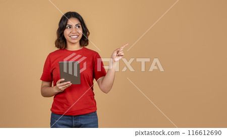 A young middle eastern woman, wearing a red t-shirt and blue jeans, is holding a tablet and pointing her right hand to copy space while smiling. She is standing against a light brown background. 116612690