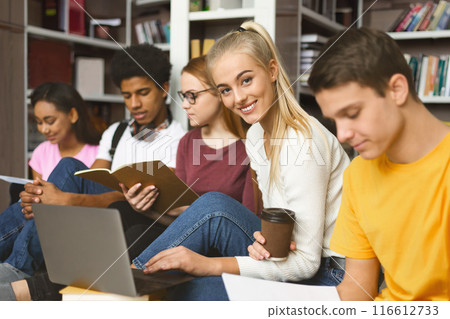 Focused international students sitting on wooden floor at library, getting ready for exams with books and laptops 116612733