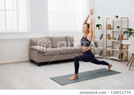 A young woman in athletic wear practices yoga in a bright living room with white walls, hardwood floors, and a couch. She is holding a warrior one pose with her arms raised overhead. A young woman in athletic wear practices yoga in a bright living room with white walls, hardwood floors, and a couch. She is holding a warrior one pose with her arms raised overhead. 116612739