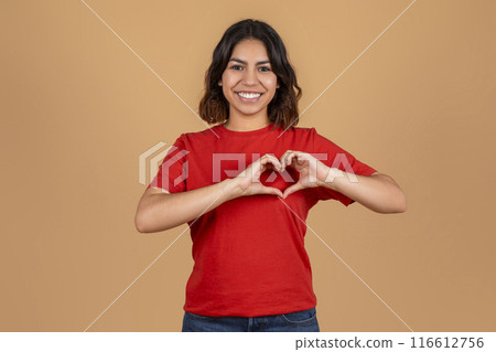 A young middle eastern woman with brown hair and a warm smile, is wearing a red t-shirt and making a heart shape with her hands in front of a light brown background. A young middle eastern woman with brown hair and a warm smile, is wearing a red t-shirt and making a heart shape with her hands in front of a light brown background. 116612756