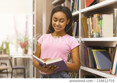 Smiling african american girl student reading book between bookshelves in campus library, empty space Smiling african american girl student reading book between bookshelves in campus library, empty space 116612759