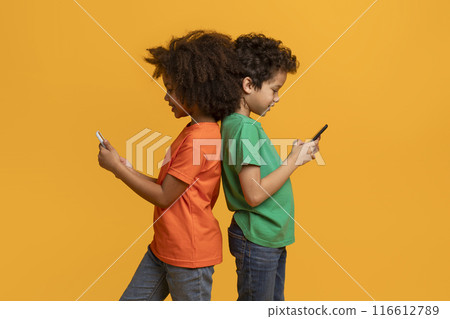 This image shows two African American children standing back to back in front of a yellow background. They both have their heads bent down, looking at their smartphones. 116612789