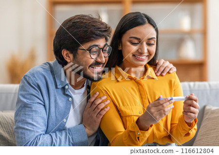 A joyful Indian couple is sitting together on their couch, delightedly examining a positive pregnancy test result in their living room during the daytime. 116612804