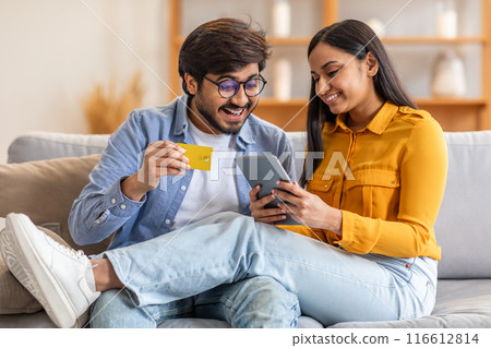 A cheerful Indian couple sits closely on a comfortable couch, engaging in online shopping using a tablet and credit card in their living room, smiling and relaxed, enjoying their time together. A cheerful Indian couple sits closely on a comfortable couch, engaging in online shopping using a tablet and credit card in their living room, smiling and relaxed, enjoying their time together. 116612814