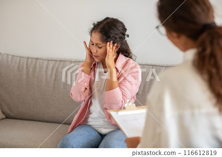 A woman sits on a couch, her head in her hands, appearing anxious during a therapy session. The therapist sits beside her, looking concerned. 116612819