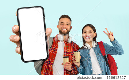 This image features two young, happy students, a man and a woman, holding coffee cups and a smartphone with white screen. They are standing against a blue background and smiling for the camera 116612827
