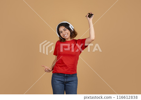 A young middle eastern woman is seen dancing and smiling while wearing white headphones and holding a smartphone in her right hand. 116612838
