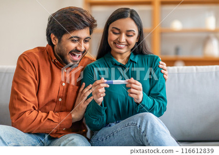 A joyful Indian couple sits on a sofa in their cozy living room, both smiling and looking excitedly at a positive pregnancy test. 116612839